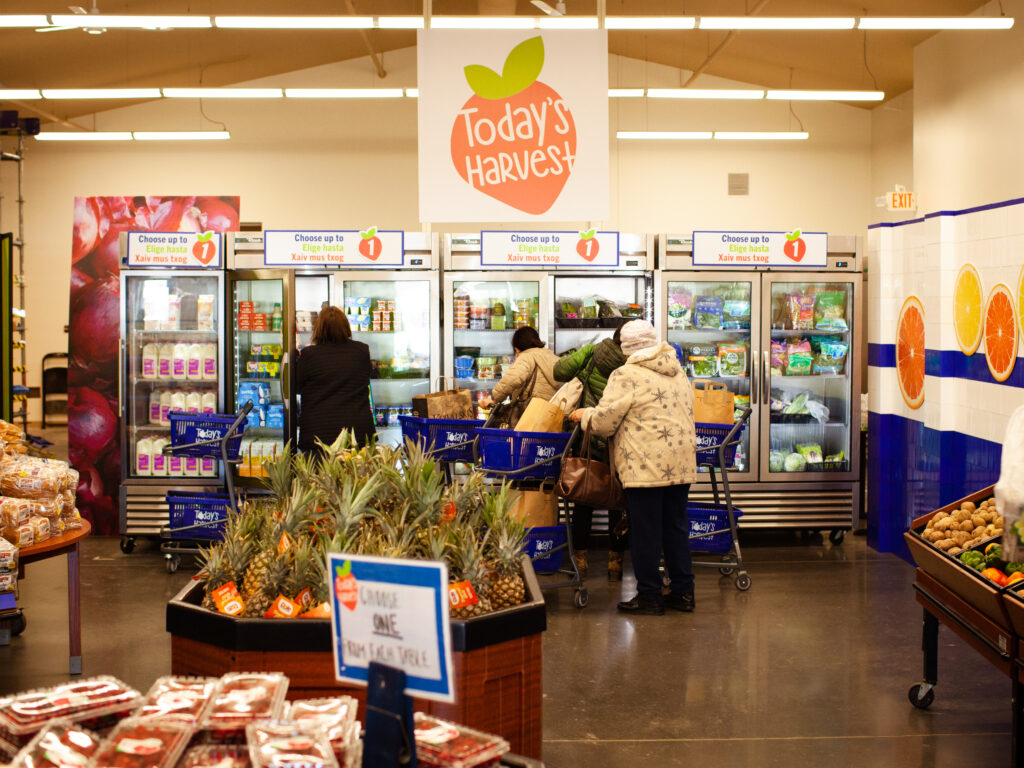 A shot of the Today's Harvest market. In the background are shoppers looking at coolers for items, in the foreground are bins of produce. A Today's Harvest logo is centered over the shoppers.