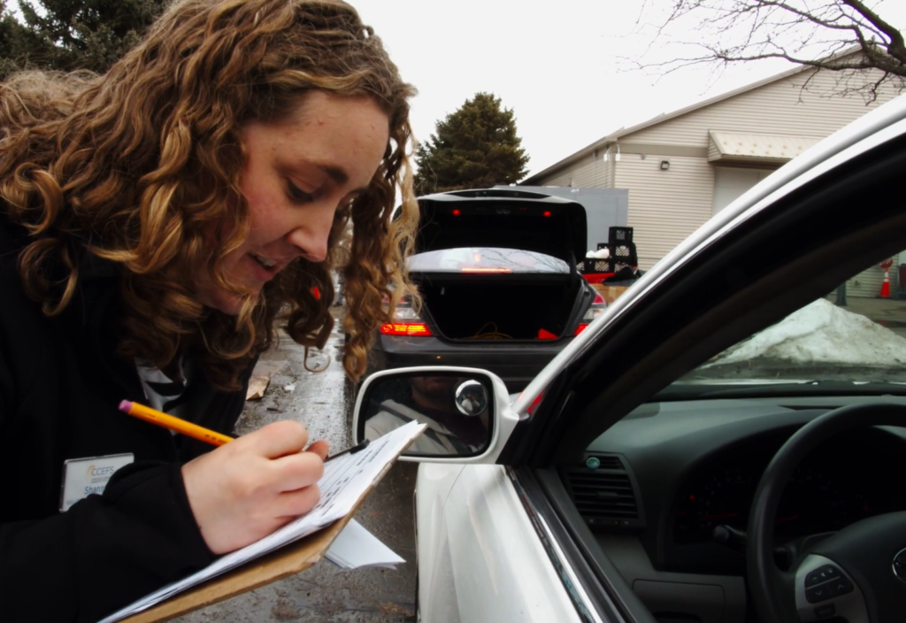 A woman with curly hair is leaning towards a car window with a clipboard, writing down information